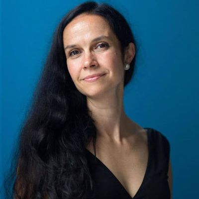 Portrait of a woman with long dark hair wearing a sleeveless black top, standing against a bright blue background.